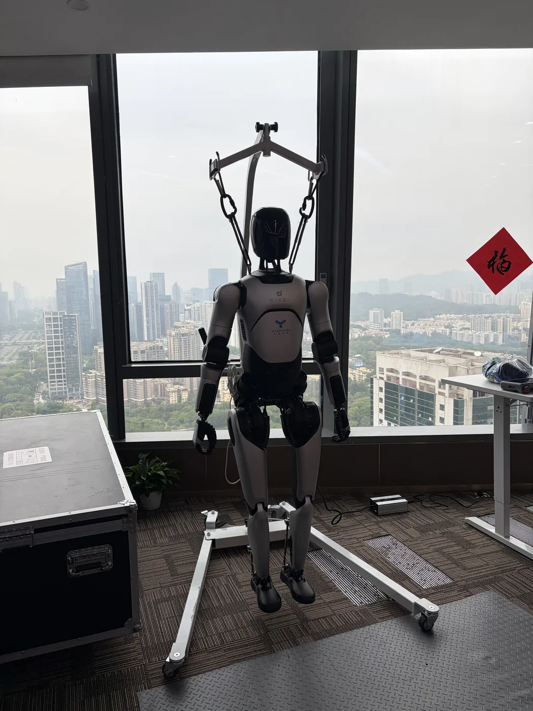 Humanoid robot suspended in a test harness, Shenzhen skyline in the background